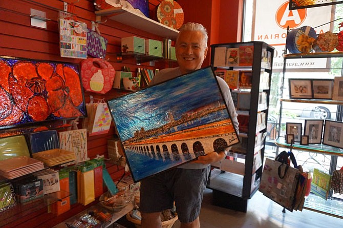 Artist Eric Nelson holds an original piece depicting the skyline of Alexandria from across the Woodrow Wilson Bridge. After five years in the Carlyle area, the Artfully Chocolate Bistro owner will be closing his doors May 27 and selling dozens of his original works through Alexandria-based online auction house Rasmus Auctions.
