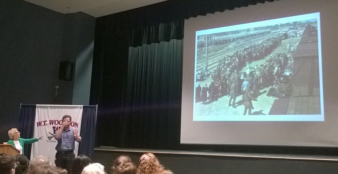 Fairfax County resident Irene Weiss, left, points to an enlarged photograph of Hungarian Jews while they were selected and processed in 1944 by Schutzstaffel guards as they stepped off a train at the Auschwitz-Birkenau concentration camp in Nazi-occupied Poland.  At the time, Weiss was the young woman with a kerchief — shown at the bottom of the picture, second from the left.  Her mother gave her the scarf to cover the teen’s shaved head.  It saved her life because it made Weiss appear old enough to join the ranks of forced laborers.  People too frail to work plus women and their children are shown at the top of the page, from left to right, walking toward the gas chamber.  Weiss was just separated from her younger sister and watched the child disappear into the crowd.  “The drama of the separation still lingers with me today,” she said April 11 to 10th-grade students at W.T. Woodson High School in Fairfax.
