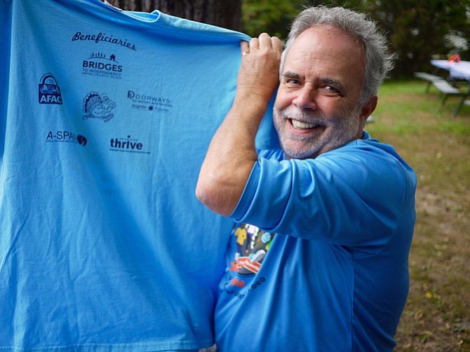 Mark Riley holds up the Turkey Trot T-shirt sponsor list on the back, pointing to the empty spaces where sponsors are still pending. Riley notes that he needs sponsors, donations, volunteers, and people to register for the race. 
