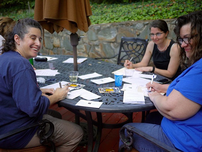 (l-r) Rebecca Heaney, Susan Cunningham, and Dana Milburn sit at the table writing postcards on Liz Lord’s patio.
