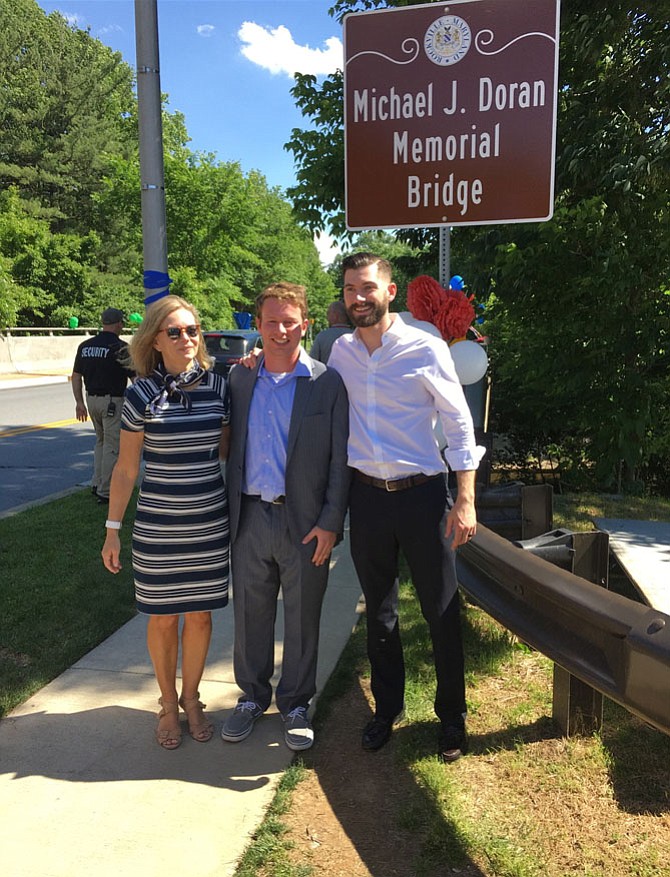 Kathy Lavinder Doran, widow of Dr. Michael Doran, with their son Sam Doran, left, stand with Jon Glaser in front of the sign designating the bridge on Wootton Parkway below Thomas S. Wootton High School in Rockville as a memorial to Doran who served as principal at the school until his death in 2015. Glaser, who graduated from Wootton in 2015, was instrumental in getting the bridge named for Doran.
