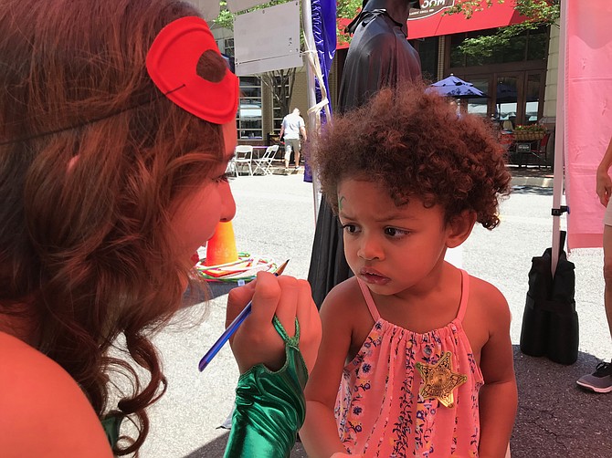 Lucia Drake, 2 1/2, seems a little shy about having her face painted by Faith Carlson, member of the Vienna Singing Princesses at Taste of Reston 2018.