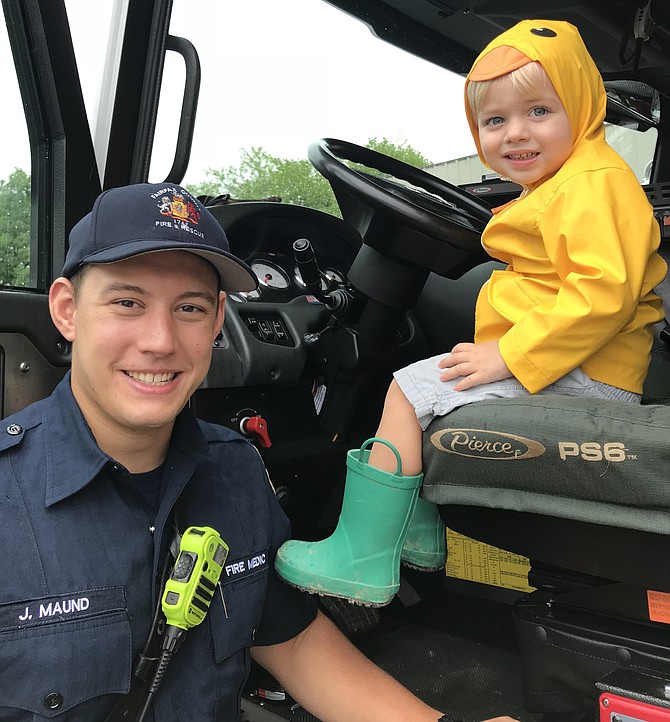 Fire Medic Jake Maund of Fairfax County Fire and Rescue, #425 Reston spots Beckett Wilson, 2 1/2 of Fairfax, as he sits in one of the County’s fire engines and tries out the steering wheel during Reston Association's annual "Totally Trucks" event held this year on Friday, Aug. 3, 2018.