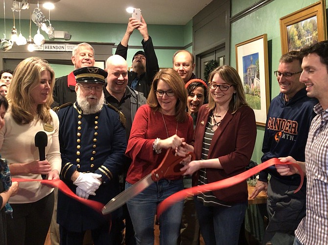 Officially opening historic Herndon's newest business, Elden Street Tea Shop, (center) Mayor Lisa C. Merkel and Rachel Eisenfeld cut the ceremonial ribbon while (far left) Vice Mayor Jennifer Baker, (second row) Town Councilmember Bill McKenna, Del. Jennifer Boysko (D-86) and District 33 Senate candidate, (third row left) Mike O-Reilly, former mayor of Herndon and current member of the Dulles Chamber of Commerce and John Boylan, President and CEO of the Dulles Chamber of Commerce cheer them on.