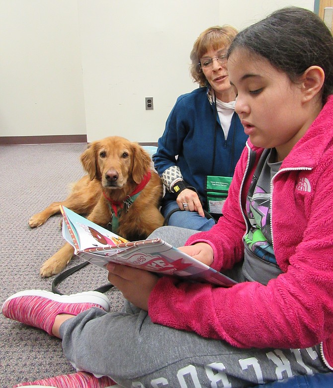 Ava Robertson, 9, of Herndon, reads her book, “Everyone Loves Cupcake,” to Copper and his handler Julie Galton of Oakton, during Dog Tales, a reading program at Herndon Fortnightly Library.