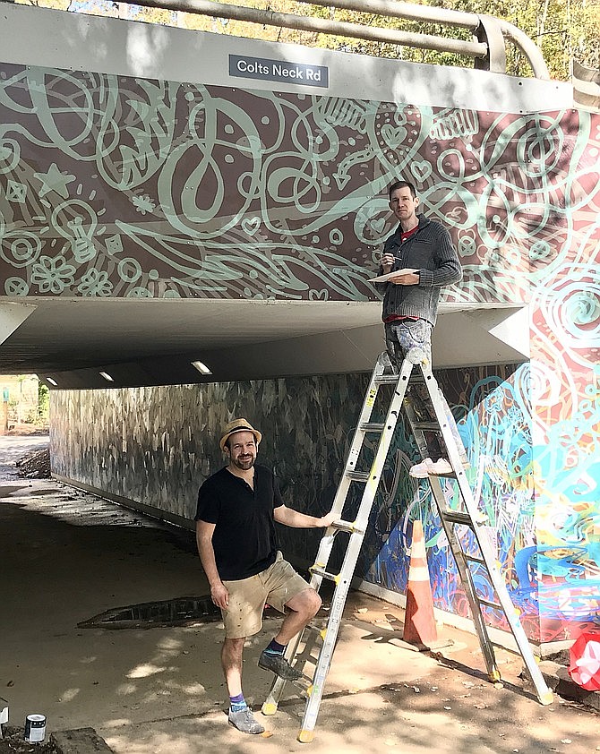Artist Ben Volta talks with James Tafel Shuster as he paints panel edges to ensure color consistency in the massive Colts Neck Road underpass mural, a commissioned artwork in Reston. (File photo)