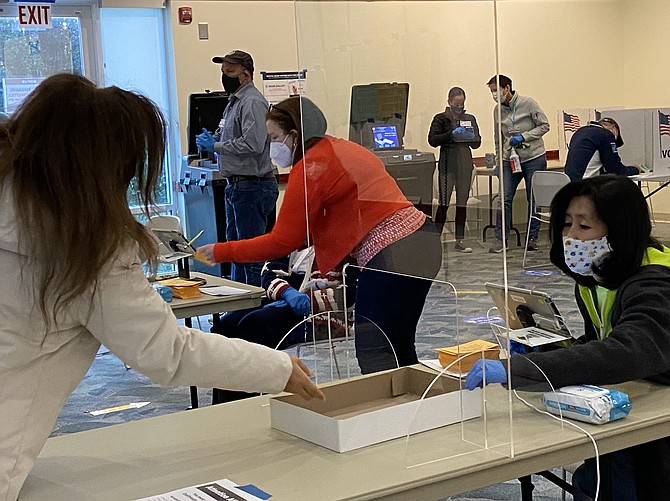 Separated by a plexiglass shield, Fairfax County election official Elizabeth Yang (far right) reaches toward the no-contact box to check the identification of voter at the Great Falls Library polling place.