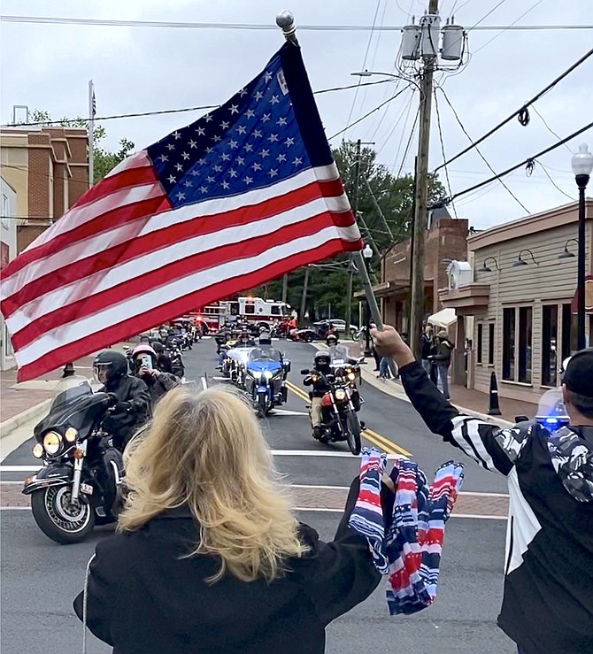 Three members of the Town of Herndon Police Motorcycle Squad lead the Jimmy’s Tavern Memorial Day motorcycle convoy down Spring Street, turning onto Elden Street where Town Mayor Sheila Olem waves her red, white, and blue scarf, and Joshua Jay waves a large United States flag bidding them a safe journey to Washington, D.C. for the Rolling to Remember ride 2021.