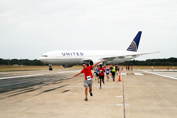 A runner poses his arms like airplane wings as he leads a pack of runners past a widebody United Airlines aircraft.