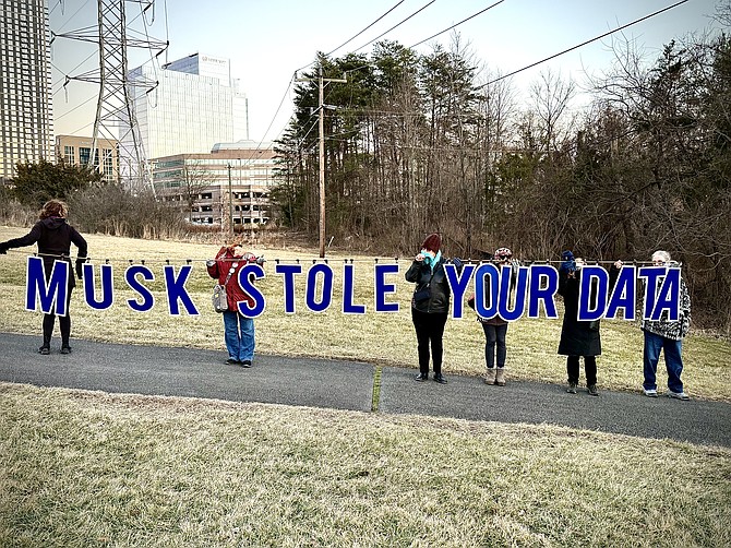 Fifteen members of the local group, Defending Democracy Indivisibles, hold letters spelling out their message of the day, “Musk Stole Your Data.”