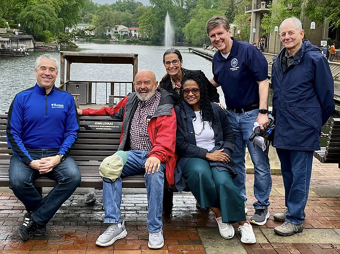 John Lovaas (second from left) tries out the bench at Lake Anne in Reston that includes a plaque honoring him for his contributions to the community, including founding the Reston Farmers Market. From left, Max Cummins, CEO of Reston Association, John Lovaas, honoree, Sen. Jennifer Boysko, Del. Karen Keys-Gamarra, Supervisor Walter Alcorn, and Irwin Flashman, RA board, Lake Anne director.