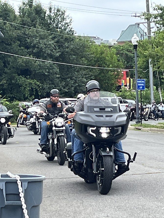 A large motorcycle group passing by the HerndonPRIDE event enthusiastically waves and gives event goers thumbs up, creating a sense of community between them.