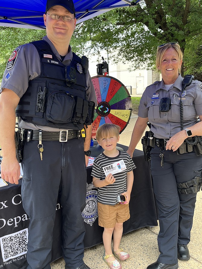 Ezra Spielvogel, 4 of Reston, beams with joy as he stands next to MPO Derek Brown, the Crime Prevention Officer, and MPO Jenette Post, the Community Outreach Officer, both from the Reston District Station. The trio posed after Ezra spun the Fairfax County Police Department’s prize wheel and won a Slurpee at the National Night Out 2025 event held at Hunters Woods in Reston.